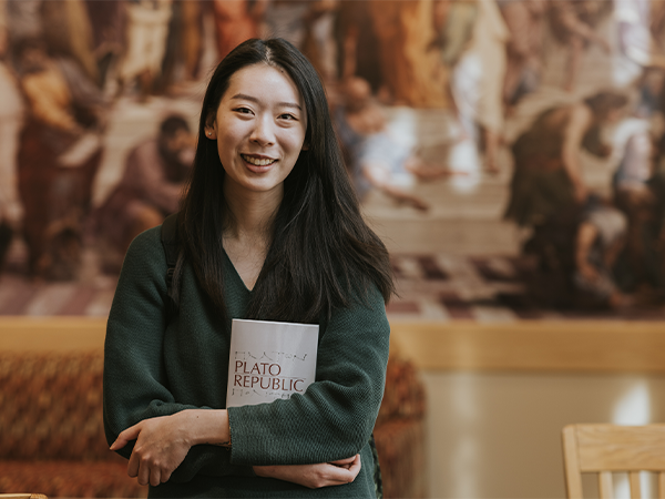 A student stands in front of a classical mural, clutching a book.